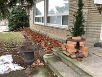 Two cast iron urns outside next to a house, one urn is intact on the ground, the other urn is placed on a set of concrete stairs.