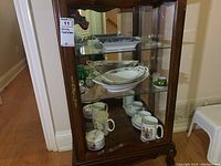 Full view of wooden china cabinet with three shelves filled with mugs, platters, cups, and vases.