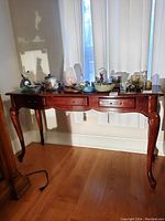 Front view of a brown wooden console table with a drawer missing handles, minor scratches visible, standing on a wood floor in front of vertical blinds.