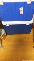 Two blue rectangular coolers stacked, white latches, placed on wooden floor.