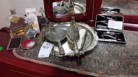 Photo showing silver-tone decorative tray holding handheld mirror, brush, comb, and a decorative glass cologne bottle with stopper, along with additional partial cologne bottles and grooming kit in case on patterned fabric dresser cover beneath mirror.