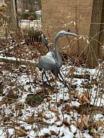 Two metal heron sculptures positioned outdoors in a garden bed with dead grass and snow patches visible. The herons are gray and approximately 3 feet tall with surface wear from weather exposure.