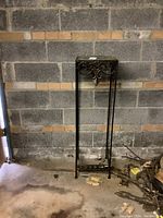 Full view of the tall metal plant stand against a block wall, showing the overall form and structure with a glass surface on top.