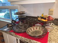 Wide view of the kitchen counter displaying all silver plated items: stack of cake stands, fruit bowl with faux fruit, cups, baskets, and trays on red cloth.