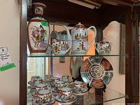 Front view of entire tea set displayed on glass shelf inside wooden cabinet, showing vase, teapot, sugar bowl, creamer, 10 matching teacups with saucers, and decorative plate.