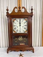 Front view of hardwood table clock with colorful floral design on clock face, roman numerals, and decorative gold design on glass door. Clock key is shown in front of the clock.