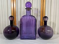 Front view of three purple glass containers: one large rectangular shape in center, two smaller round shapes on either side, all with stoppers