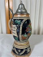 Full view of the beer stein showing the detailed ceramic body with relief artwork and metal lid.