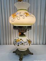Front view of vintage hurricane glass lamp showing white iridescent glass with hand painted yellow and blue flowers and decorative metal base.