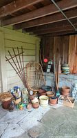 Wide view showing hanging bamboo chair, garden tools in baskets and pots, numerous planter pots, and garden art figures including Sphinx bust and Pan figure.
