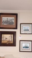 Photo showing four framed artworks hanging on a wall: top left is a Ghirardelli city scene in black and white, bottom left is a pheasant painting, right side includes two sailing ship paintings by Jack Spurling.