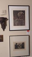 Photo of wall showing framed artwork 'Atlantis' and 'The Pardee House' along with ceramic monk shelf and owl tile.