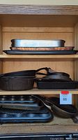 Shelves with stacked baking trays, cookie sheets, and steel baking pans of various sizes on the top shelf. Cast iron pans and pots including a skillet beneath. Muffin tins and a cast iron pan on the bottom shelf.