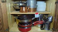 Lower shelf showing red frying pans stacked, metal colander, stainless steel pot and sifters