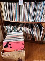 Photo showing two wooden shelves about 34.5 inches wide filled with LP records and one box containing about 95 LPs mainly musicals and soundtracks.