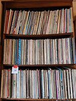 Three wooden shelves filled with assorted vinyl record LPs, organized side by side.