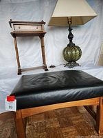 Photo showing leather and wood stool in front, green glass lamp with off-white shade and small wood phone table in background.