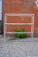 Full front view of aluminum square tube frame shelf against a brick wall, showing its rectangular shape with three horizontal shelf levels.