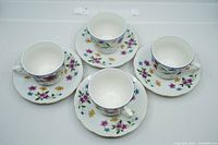 Four white china teacups and four matching saucers arranged in a group, showing floral pattern with multiple colored flowers and gold trim on rims.