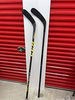 Two left handed hockey sticks, CCM Tacks 405 and Sher-Wood Rekker Euro, standing upright against white panel and red roll door.
