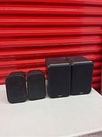 Front view of two pairs of black speakers on white surface against a red background, showing size and appearance.