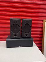 Two B&W bookshelf speakers and one Sound Dynamics center channel speaker placed on table with red background.