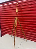Full view of a wooden adjustable easel against a red metal storage background on a light surface.