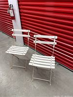 Two antique white metal and wood fold-up patio chairs in front of red corrugated wall.