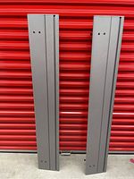 Two heavy duty metal narrow shelves standing vertically against a red corrugated metal background. Shelves show grooves running lengthwise and holes possibly for attachments.