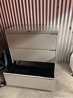 Gray metal horizontal filing cabinet with three drawers, one drawer open showing black interior and file rails. Located indoors on concrete floor with corrugated metal wall background.