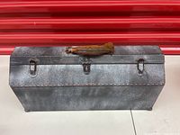 Front view of the rectangular metal toolbox showing rusted handle and latch hardware on top surface against a red background.