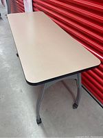 Side angle photo showing rectangular beige desk top with black edge and curved metal legs with caster wheels.