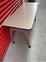Full view of the rectangular desk/table showing beige laminate top with black edges, metallic legs, and caster wheels.