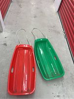 Two plastic snow sleds placed side by side on a concrete floor in front of red storage unit doors. One sled is bright red and the other is green. Both have white pull ropes and side handles visible.
