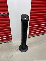 Full height image of black Seville Classics tower fan on round base against red storage door background.