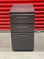 Front view of gray metal filing cabinet with three closed drawers showing locking key on top drawer.