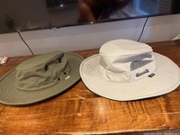 Two Tilley hats side by side on a wooden surface: one khaki, one beige, showing their full profile and brims.