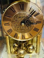Close-up of the gold-tone anniversary clock face with black ornate hands, Roman numerals, brand "ergq", and decorative swirls.