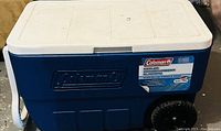 Side view of the blue and white Coleman cooler with wheels and handle, showing the Coleman branding and label.