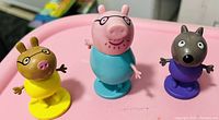 Photo of three Peppa Pig character figurines on a pink surface.