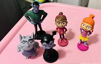 Five colorful plastic figurines from Vampirina arranged on a pink surface, showing all characters.