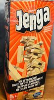 Box of the Classic Jenga game showing the logo, artwork, and branding details including Hasbro branding and age recommendation of 6+. The box art depicts wooden blocks being stacked in a game setup.