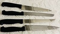 Photo shows four kitchen knives with different blade shapes and black handles, laid out on a textured white surface.