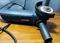 Blue Mastercraft electric angle grinder shown from the side with visible grinding disc and attached side handle, corded power design.