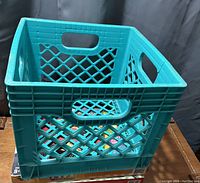 Teal plastic milk crate with lattice sides, handle cutouts on three visible sides, sitting on table with some board games beneath.
