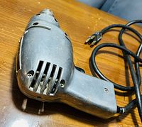 Photo of a metal-bodied corded electric power drill on a wooden surface, showing the front with ventilation slots and attached black power cord with plug.