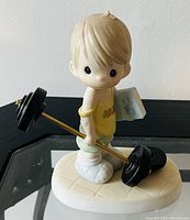 Front view of the Precious Moments figurine showing the child holding a barbell and book, standing on a tiled base.