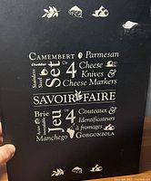 Photo of black box with white typographic text and small cheese related illustrations describing the cheese knife set's contents and brand 'Savoir Faire'