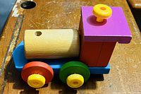 Photo showing a wooden toy train with a natural wood cylindrical smokestack, a purple square cab with a yellow knob, blue base, and four colorful wheels in red, green, and yellow.