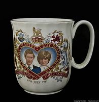 Front side of white ceramic mug with portraits of Charles and Diana inside a heart-shaped frame decorated with royal heraldry and the date 29th July 1981.